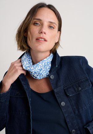 Sieviete ar īsiem brūniem matiem, valkājot rakstainu zili baltu kaklasaiti, tumši zilu jaku un melnu topu pret vienkāršu fonu.