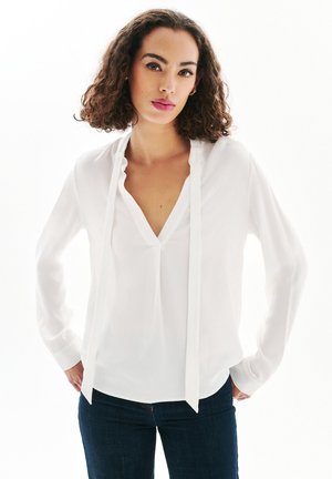Blusa blanca de mangas largas con escote en V profundo y detalle de lazo en el frente, hecha de un tejido suave y ligero. Combinada con jeans azul oscuro.