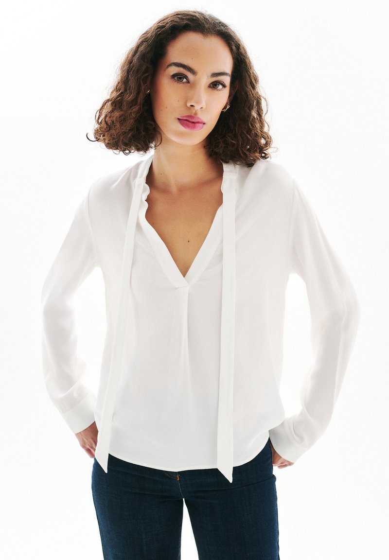 Blusa blanca de mangas largas con escote en V profundo y detalle de lazo en el frente, hecha de un tejido suave y ligero. Combinada con jeans azul oscuro.