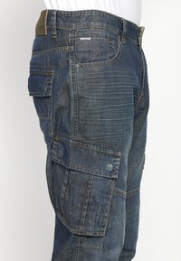 Sötétkék denim cargo nadrág, több nagy zsebbel, megerősített varratokkal, és texturált felülettel, amelyen látható varrási részletek találhatók.