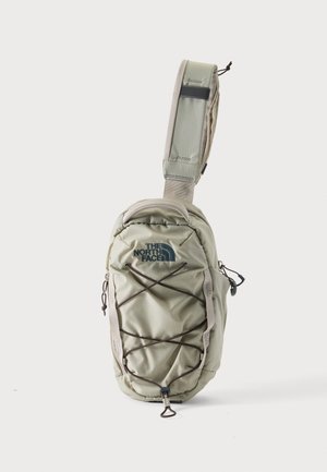 Lysegrå North Face sling taske med polstret rem, elastisk snor foran og lynlåslommer på hvid baggrund.