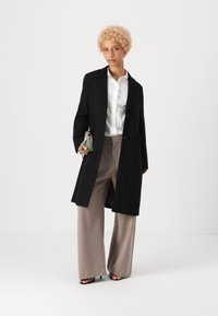 Cappotto in misto lana nero con colletto a punte, chiusura a bottoni e vestibilità oversize; abbinato a pantaloni beige a gamba larga e una clutch verde testurizzata.