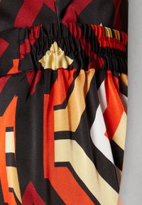 Barevná vzorovaná látka v červené, oranžové, černé a žluté s detailem na zavázaném pase. Textura vypadá hladce a lehce.