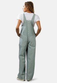 Salopette verte rayée avec jambes larges, en coton, dotée de bretelles réglables et de deux poches arrière. Portée par-dessus un t-shirt blanc.