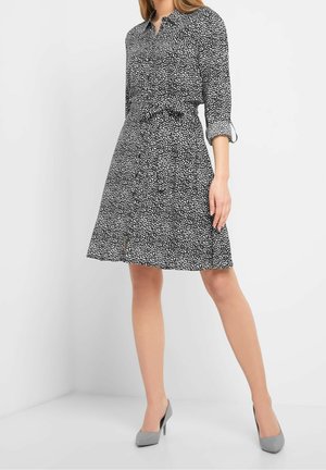 Woman wearing a black knee-length dress with white spot pattern, long sleeves rolled up, and gray high-heeled shoes standing against a white background.