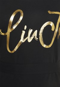 Tissu noir avec texte cursif doré brillant partiellement visible, avec les lettres visibles "lin" et une partie d'un grand "J".