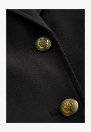 Sacou din țesătură neagră cu nasturi aurii, texturați, cu modele intricate și croială ajustată, evidențiind reverele ascuțite și liniile clare.