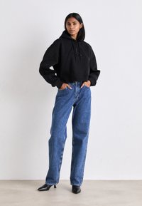 Felpa nera corta con logo, abbinata a jeans a vita alta azzurri. La modella indossa tacchi neri appuntiti, mostrando una vestibilità rilassata e uno stile casual.