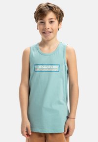Ljus teal bomullslinne med en blå och rosa grafisk logotyp. Har en klassisk rund hals och ärmlös design, i kombination med beige shorts.