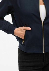 Veste bombers bleu marine avec fermeture éclair à l'avant, quincaillerie dorée, poignets côtelés et poches latérales avec fermetures éclair. Fini lisse.