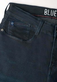 Los pantalones vaqueros de mezclilla oscura presentan un diseño clásico de cinco bolsillos con cierre de botón. La cinturilla tiene una etiqueta "BLUE" y detalles de costura en rojo que contrastan.