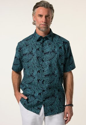 Uomo con capelli grigi e barba, indossa una camicia blu scuro a maniche corte con motivi di foglie verdi e pantaloni bianchi, in piedi con una mano in tasca.