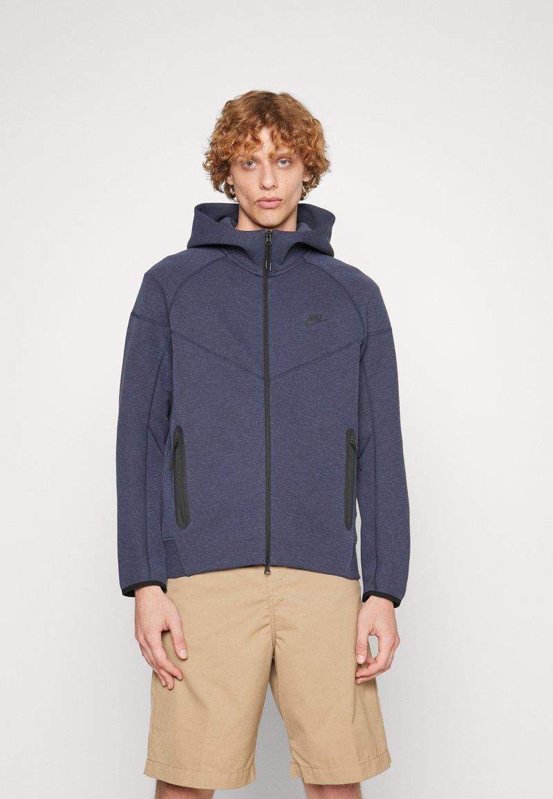 Sweat à capuche navy avec une fermeture éclair intégrale, deux poches zippées et un design à coutures courbées. Présente le logo Nike en noir.