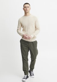 Pullover bej cu model canelat, pantaloni cargo verzi cu manșete elastice și adidași negri cu glezna înaltă. Modelul stă cu mâinile împreunate.