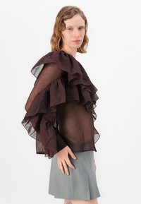 Doorzichtige bruine top met grote ruffles langs de schouders en mouwen, gecombineerd met een grijze geplooide rok. Soepele stoftextuur.