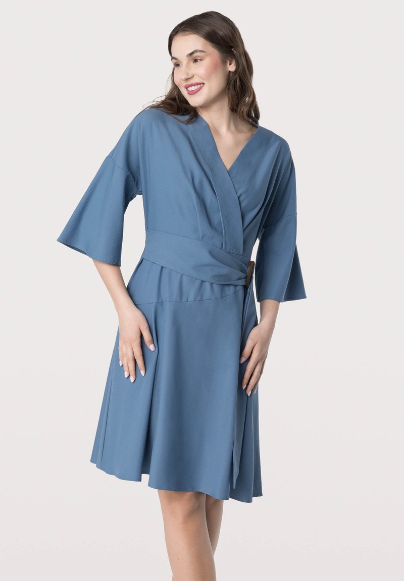 Robe portefeuille bleue avec un décolleté en V, des manches courtes évasées et une ceinture à la taille. Fabriquée dans un tissu doux, avec une jupe évasée et un détail fente sur le côté.
