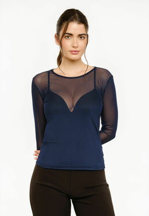 Kvinde med langt brunt hår bundet tilbage, iført en gennemsigtig navy langærmet top over en matchende camisole, kombineret med sorte bukser, stående mod hvid baggrund.