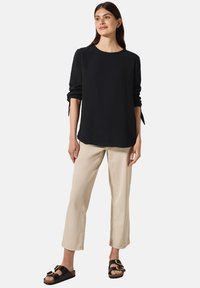 Haut noir à manches longues froissé, pantalon beige à jambe droite et sandales noires avec des détails en métal doré. Conception simple et coupe décontractée.