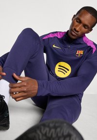 Navy athletic tracksuit with purple accents, featuring a zip-up collar and a yellow Spotify logo. Model sitting, adjusting pants, wearing black sneakers.