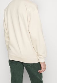 Krämfärgad sweatshirt med avslappnad passform. Slät textur, ribbad mudd och nederkant. Kombinerad med gröna denimbyxor med ett subtilt mönster.