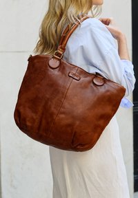 Ruda odinė tote bag su lygiu paviršiumi, suapvalintu dugnu, tvirtais dirželiais ir subtiliu logotipo akcentu. Sukurta kasdieniam naudojimui.