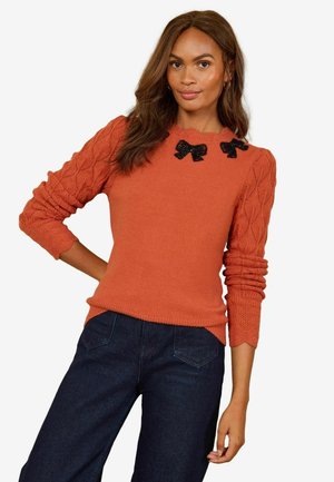 Kvinde iført en orange strikket sweater med teksturerede ærmer og to sorte sløjfedetaljer nær halsudskæringen, kombineret med mørkeblå bukser.