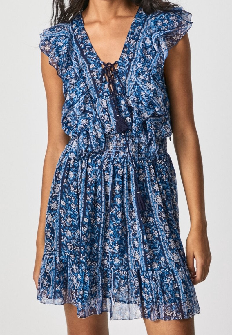 Femme portant une robe bleu fleurie sans manches, avec des épaules à volants, un col à nouer et une taille froncée.