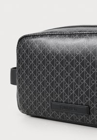 Zwarte rechthoekige tas met een gestructureerde oppervlakte, voorzien van een geometrisch patroon in donkergrijs. Leeraccenten en een merklabel van Calvin Klein zijn zichtbaar.