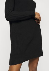 Personne portant une robe noire côtelée à manches longues, les mains posées à l'avant, avec les jambes inférieures visibles sur un fond clair.