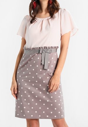 Femme portant un chemisier à manches courtes rose pâle rentré dans une jupe grise mi-longue à pois blancs avec une ceinture nouée.