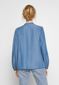 Denim shirt in light blue with long puff sleeves, button collar, and elastic cuffs. Soft texture, relaxed fit, and stitched detailing at hem.