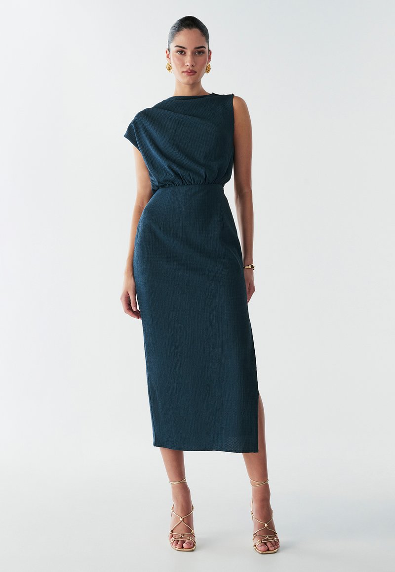 Robe midi en teal texturé avec un décolleté asymétrique, une taille froncée et une fente sur le côté. Associée à des sandales à talons dorées à lanières.
