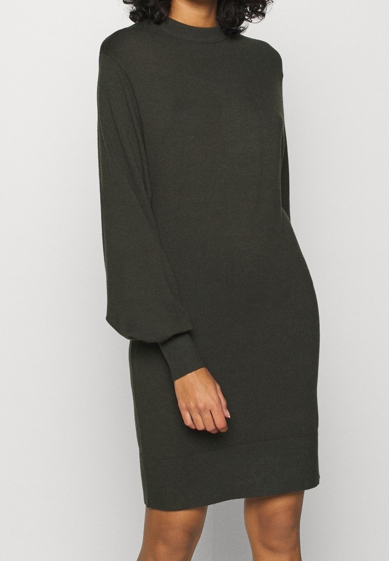 Femme portant une robe en maille vert foncé, manches longues, longueur genou, avec un col haut et des poignets ajustés.