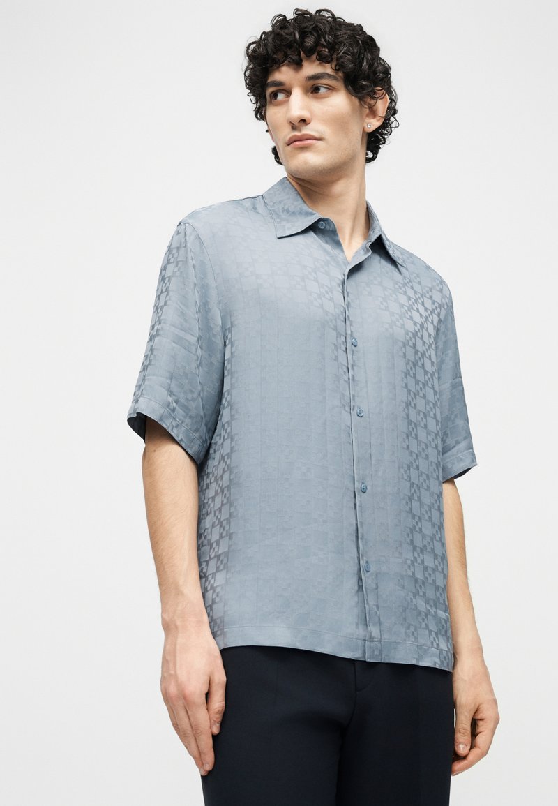 Young man with curly hair wearing a light blue short-sleeve button-up shirt with a subtle checkered pattern, standing against a plain background.