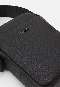 Borsa a tracolla in pelle nera con finitura texture, caratterizzata da un discreto logo stampato in rilievo e una robusta tracolla in tessuto. Forma rettangolare compatta.