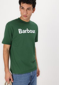 Πράσινη βαμβακερή μπλούζα με κοντό μανίκι, στρογγυλή λαιμόκοψη και χαλαρή εφαρμογή, που διαθέτει το λογότυπο "Barbour." σε λευκό χρώμα. Φοριέται με μπλε τζιν.