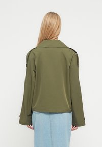 Oliivinvihreä takki, jossa on litteä kaulus ja pitkät hihat, rento istuvuus ja napit yksityiskohtina. Yhdistetty vaaleansiniseen farkkuhameeseen.