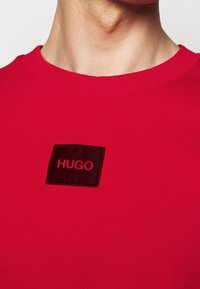 Gros plan sur une personne portant un t-shirt col rond rouge avec un patch noir centré sur la poitrine portant l'inscription « HUGO ».
