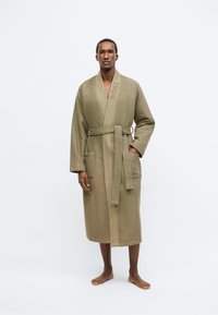Accappatoio verde oliva dalla texture a nido d'ape, con maniche lunghe, due tasche frontali, cintura da annodare e logo "BOSS" in rilievo sul colletto.