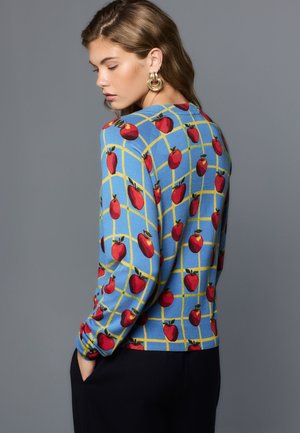 Junge Frau trägt einen blauen Pullover mit gelbem Gitter- und roten Apfelmustern, Hände in den Taschen, seitlich vor einfachem grauem Hintergrund.