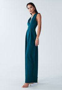 Türkisfarbener, ärmelloser Jumpsuit mit tiefem Ausschnitt, tailliertem Schnitt und weitem Bein. Kombiniert mit goldenen Riemchenabsätzen und dezenten goldenen Accessoires.