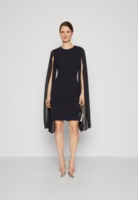 Ναυτική φόρεμα με κοντά μανίκια και ρέουσα λεπτομέρεια κάπας. Λείο ύφασμα, εφαρμοστό σιλουέτα, συνδυασμένο με ασημί ψηλοτάκουνα και μικρή τσάντα.