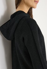 Zwarte kapjas met een gestructureerde, licht glinsterende stof. Voorzien van een nauwsluitende capuchon en raglanmouwen voor een moderne look.