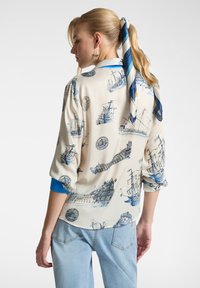Blusa bianca con design marittimi blu, che presenta navi e velieri. Dettagli blu sui maniche e una sciarpa blu annodata. Vestibilità casual.