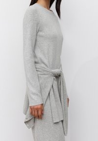 Camiseta de manga larga de color gris claro, confeccionada con tela acanalada, con un nudo anudado en la cintura y una silueta casual. Textura suave y diseño ajustado.