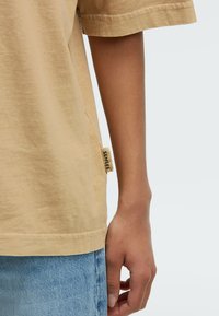 Light brown cotton t-shirt featuring a relaxed fit, stitched sleeves, and a small "SENSES" label on the side. Worn with blue denim.