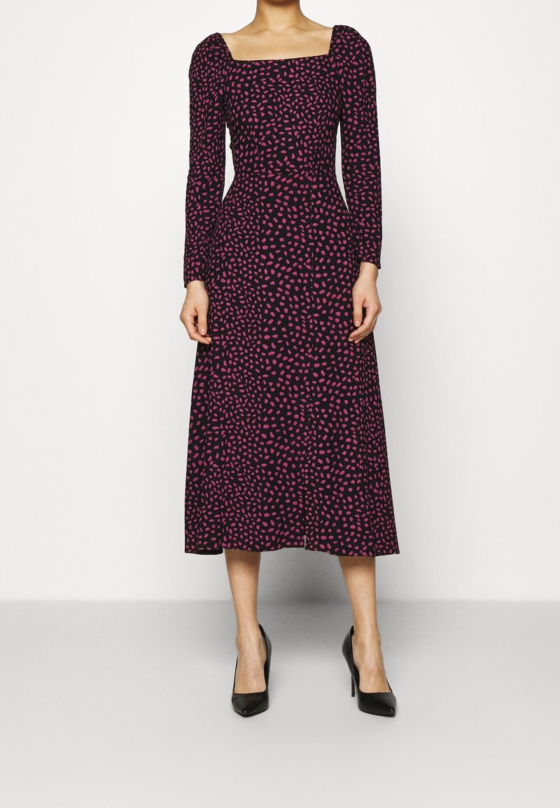 Žena v černých midi šatech s růžovými nepravidelnými tečkami, s dlouhými objemnými rukávy, čtvercovým výstřihem a černými vysokými podpatky.