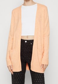 Žena v broskvovém cardigan s otevřeným předním dílem přes bílý zkrácený top a černé kalhoty s vysokým pasem a malými hvězdnými vzory.