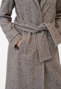 Kappa med fiskbensmönster i brunt och beige med bälte. Har en spetsig krage, fickor och subtila paljettaccenter på tyget.