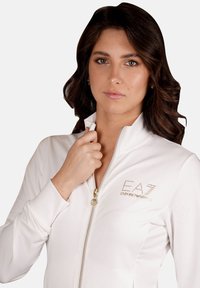 Giacca bianca con zip, caratterizzata da un alto colletto e un design aderente, con dettagli in logo dorato e una texture liscia e elastica.